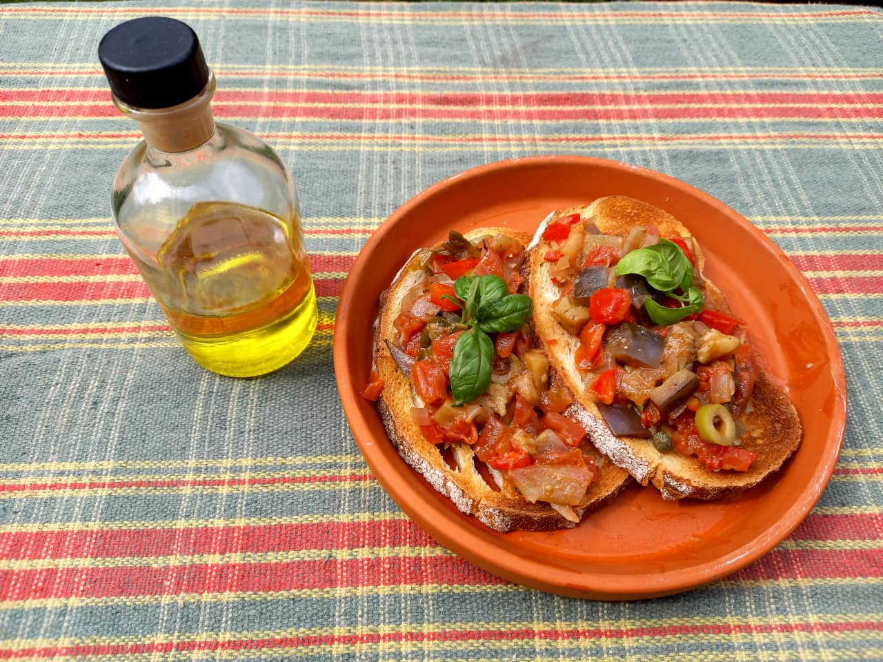 A plate with slices of bread topped with caponata next to a flask of olive oil.