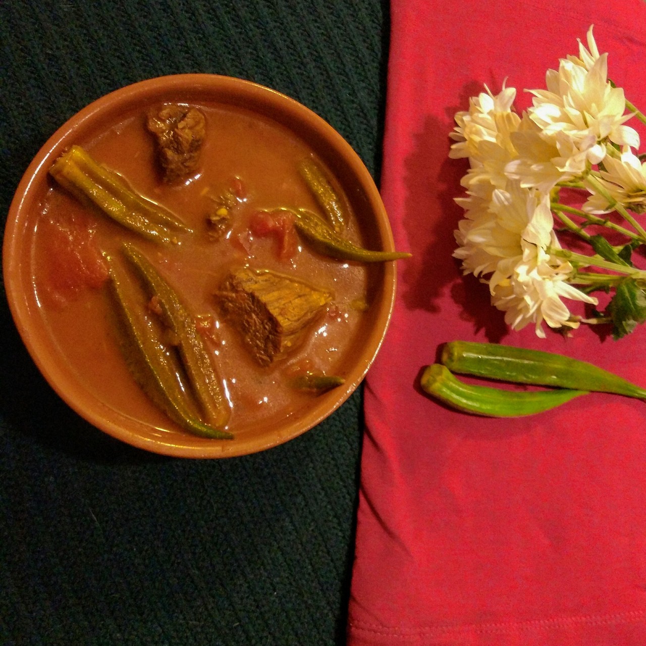 A bowl containing Khoresht Baamieh next to some white flowers and raw okra.