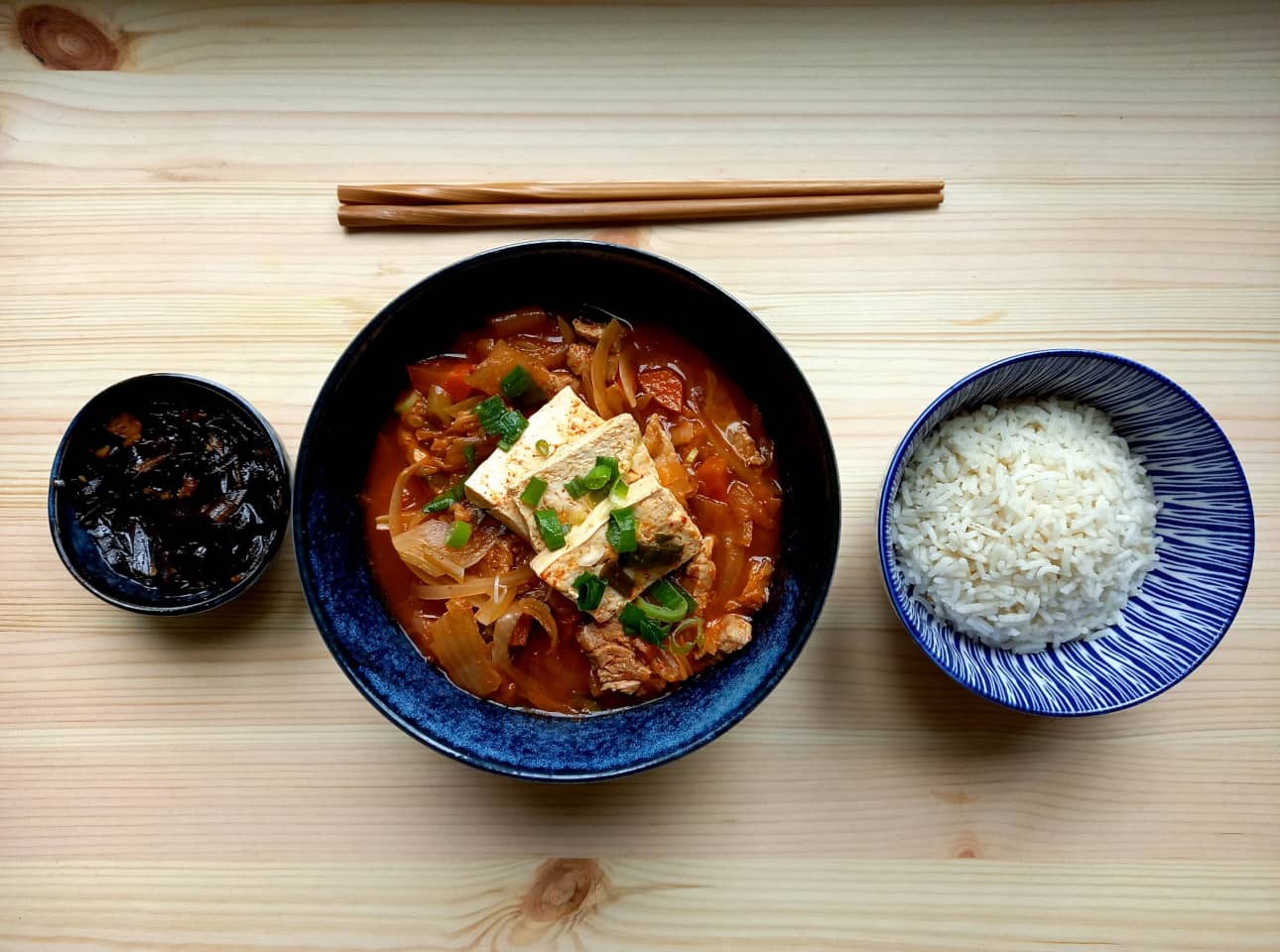 A bowl of kimchi jjigae next to a bowl with white rice and a side dish of kelp salad with soy sauce.