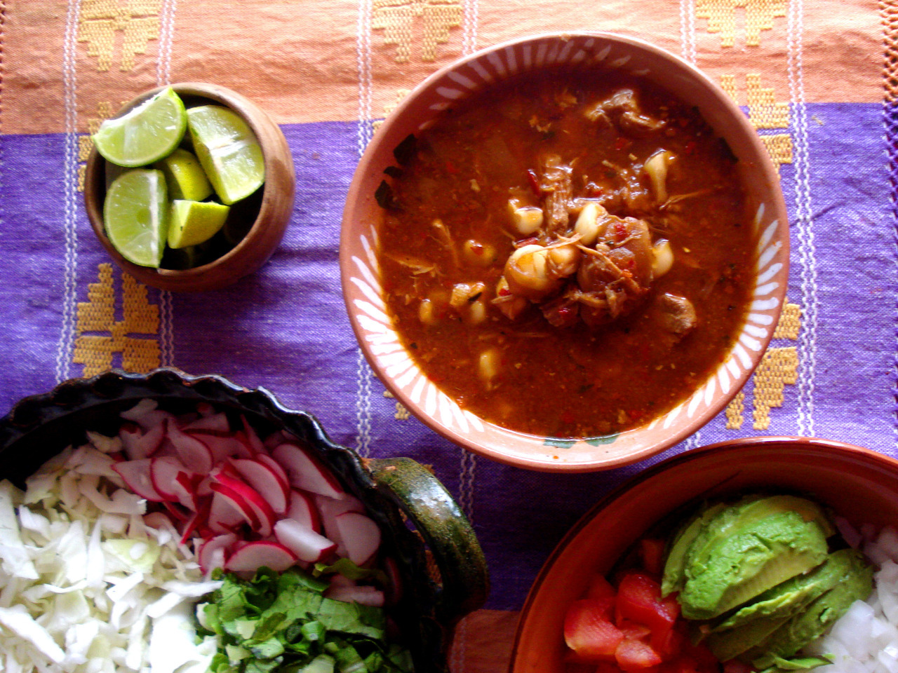 A bowl with pozole rojo next to dishes with optional additions to the stew.