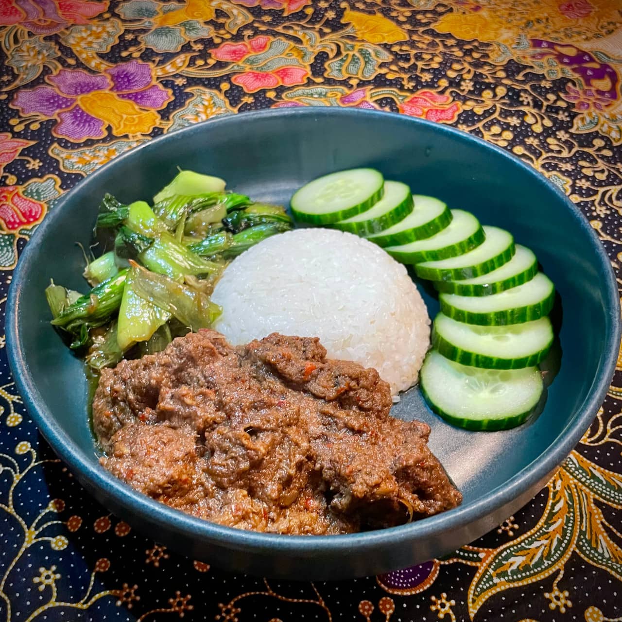 A plate of beef rendang with some stir fried bok choy and white rice, sided with cucumber slices.