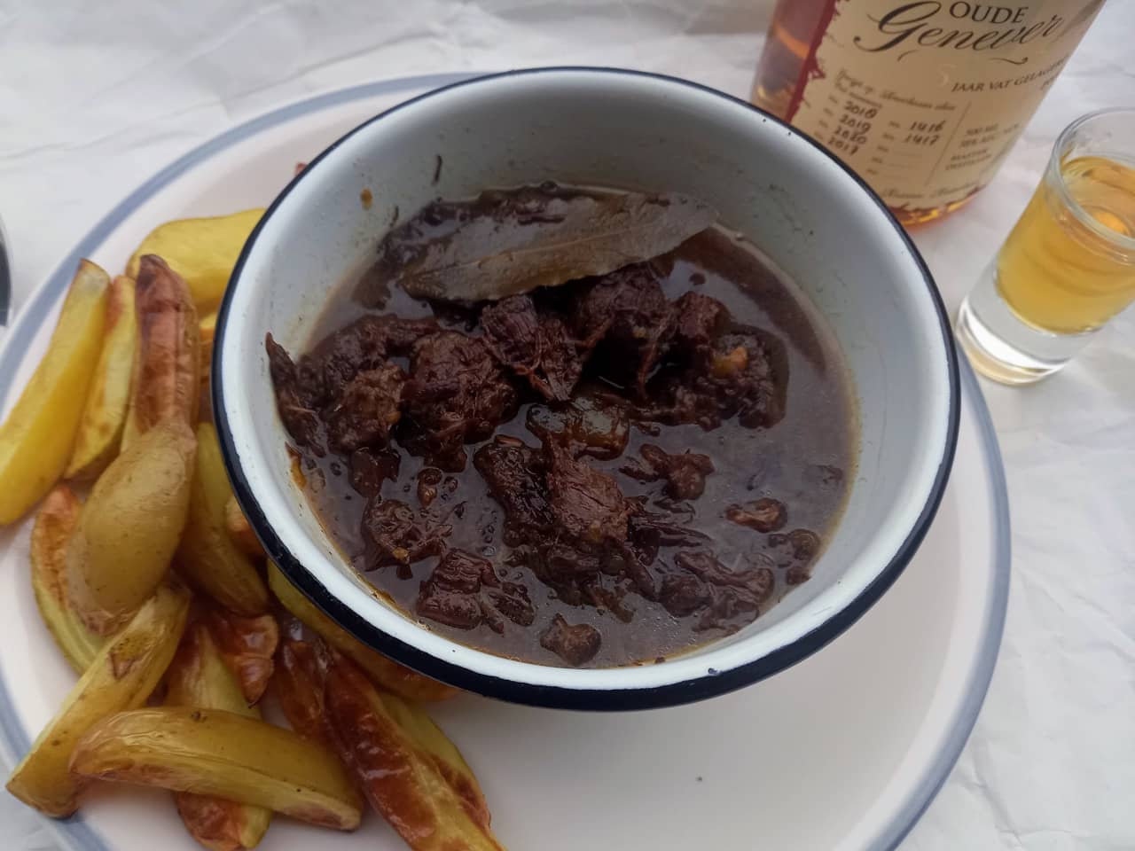 A bowl of zuurvlees and some baked potatoes next to a shot of Dutch Oude Genever.